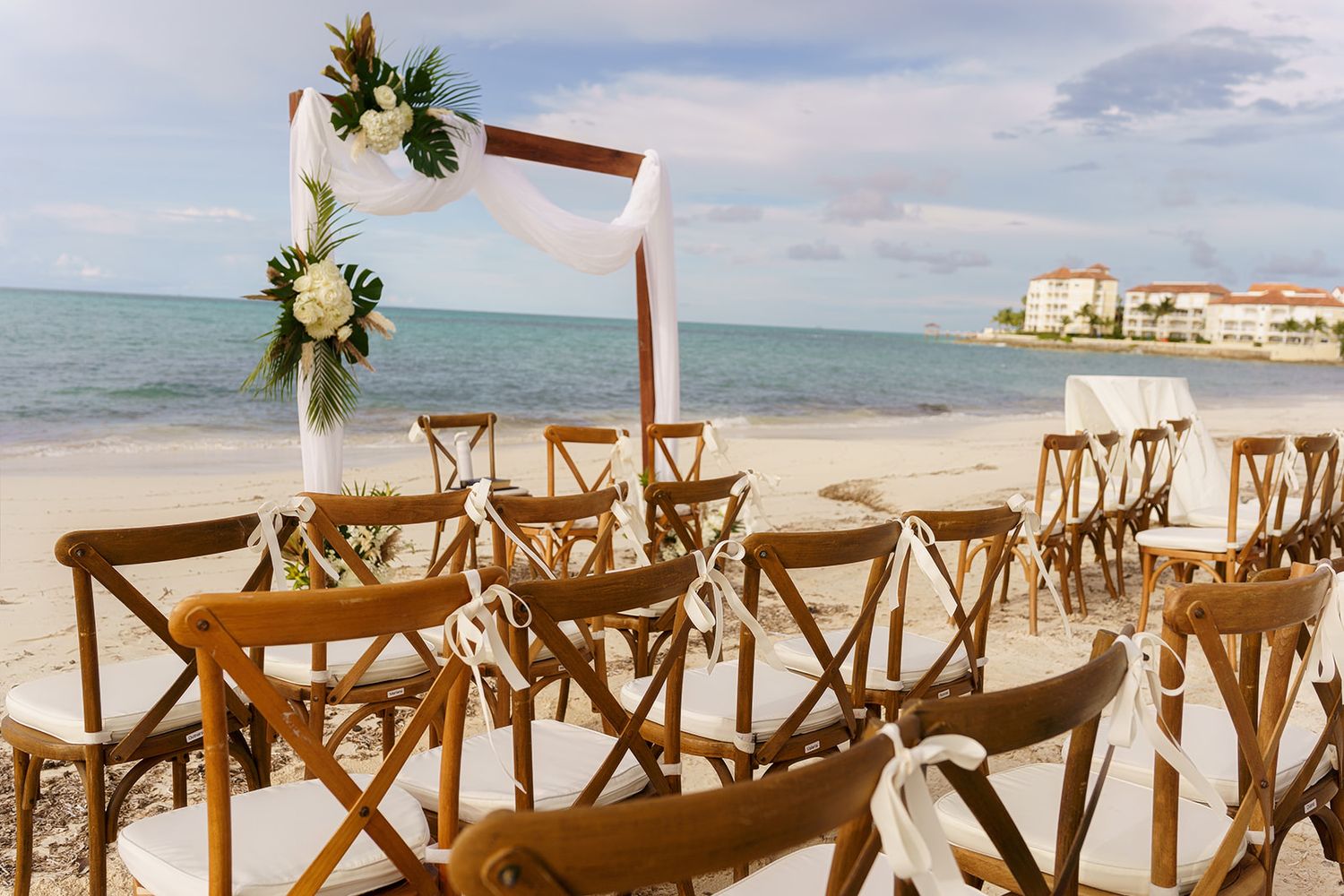 Beach wedding setup with wooden chairs and floral arch by the ocean.