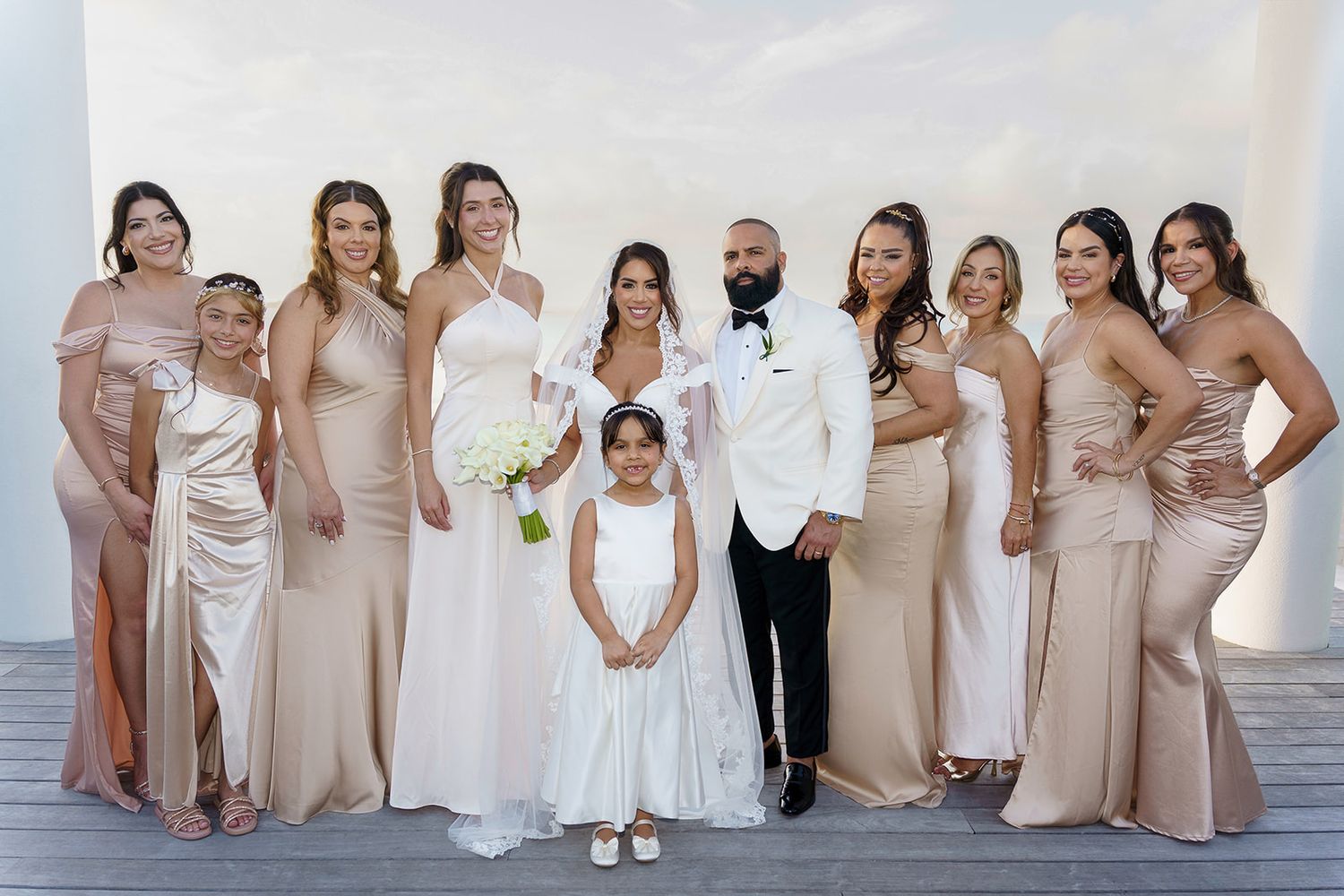 Bride and groom pose with bridesmaids in champagne gowns and flower girl on outdoor deck at wedding.