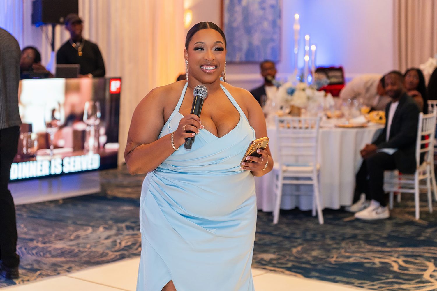Woman in light blue halter dress holding microphone at elegant banquet event.