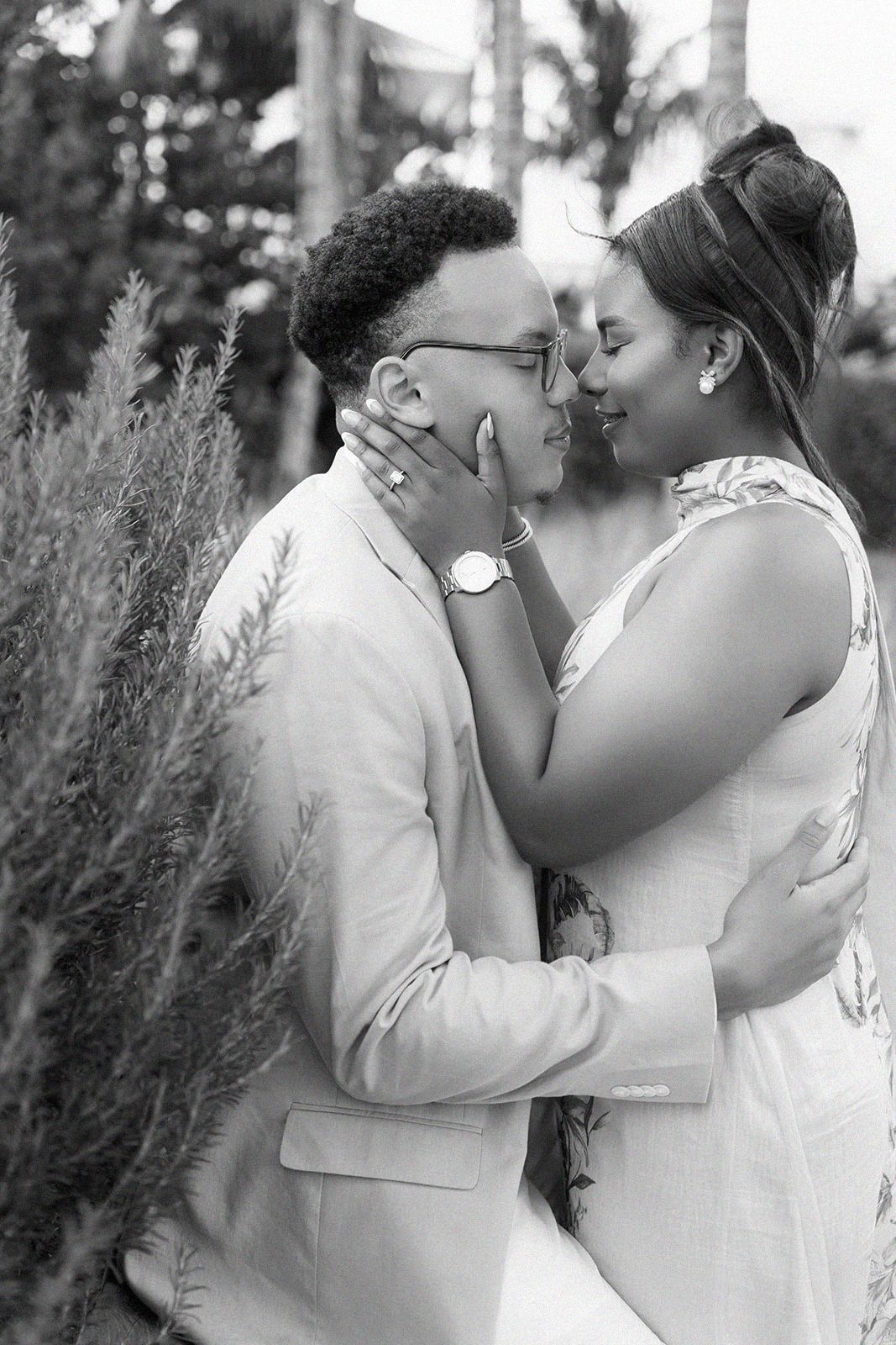 Black and white photo of a couple in formal wear sharing a romantic moment outdoors among lush greenery.