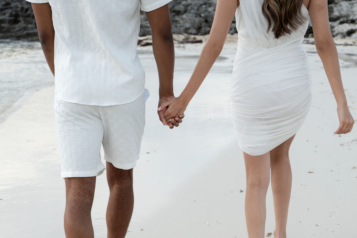 Couple holding hands walking on a white sand beach wearing matching white outfits.