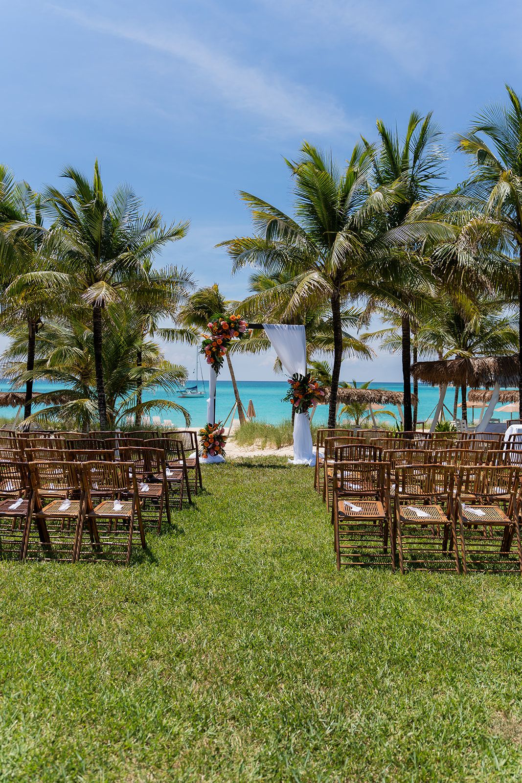 Outdoor beach wedding setup with wooden chairs, floral arch, palm trees, and turquoise ocean views under a sunny blue sky.