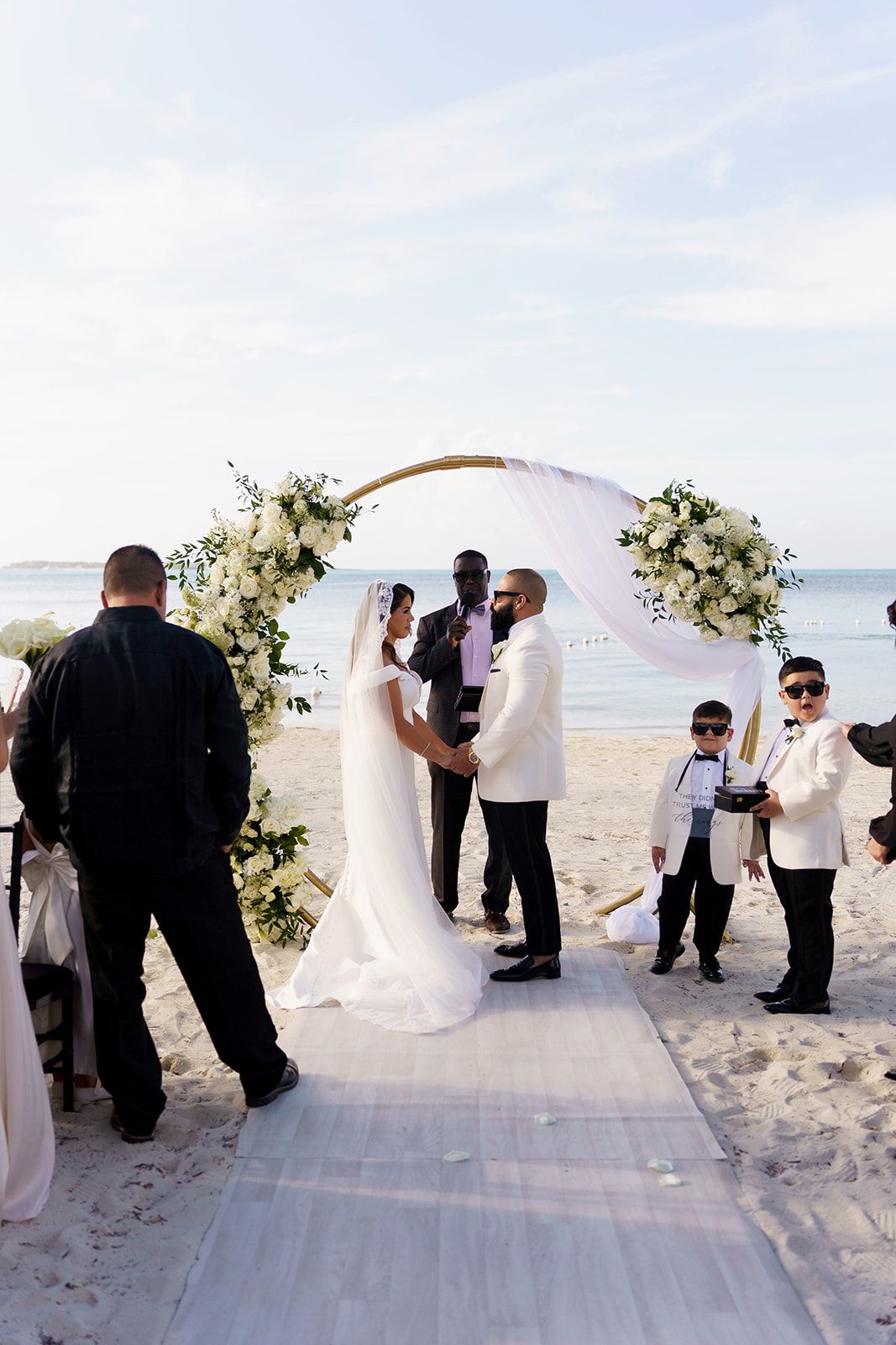 Couple exchanges vows under floral arch on beach wedding ceremony with white aisle runner and ocean backdrop.