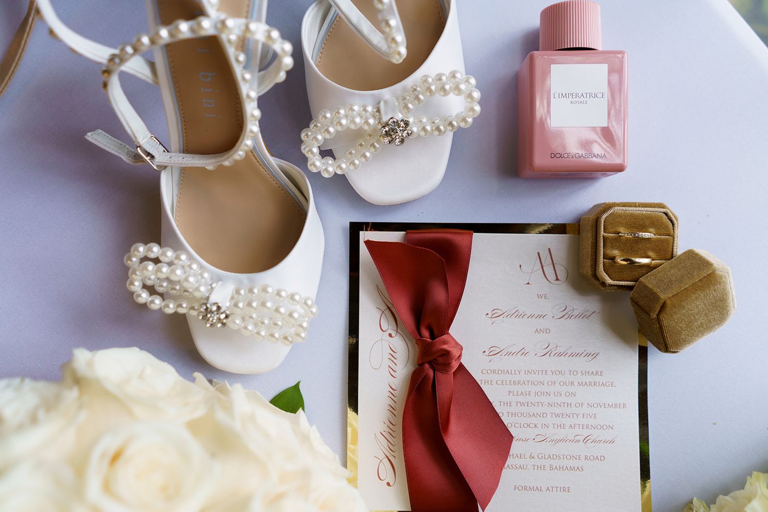 Elegant wedding flat shoes adorned with pearls beside a floral invitation with red ribbon, ring box, and pink perfume bottle.