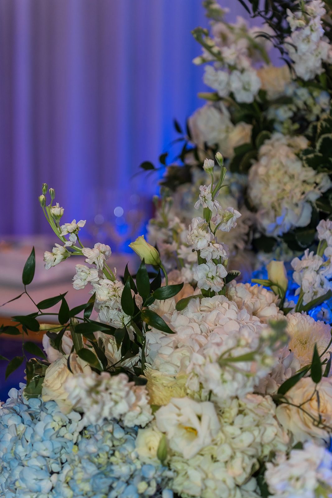 Elegant white and blue floral wedding centerpiece featuring hydrangeas, roses, and greenery in soft blue lighting.