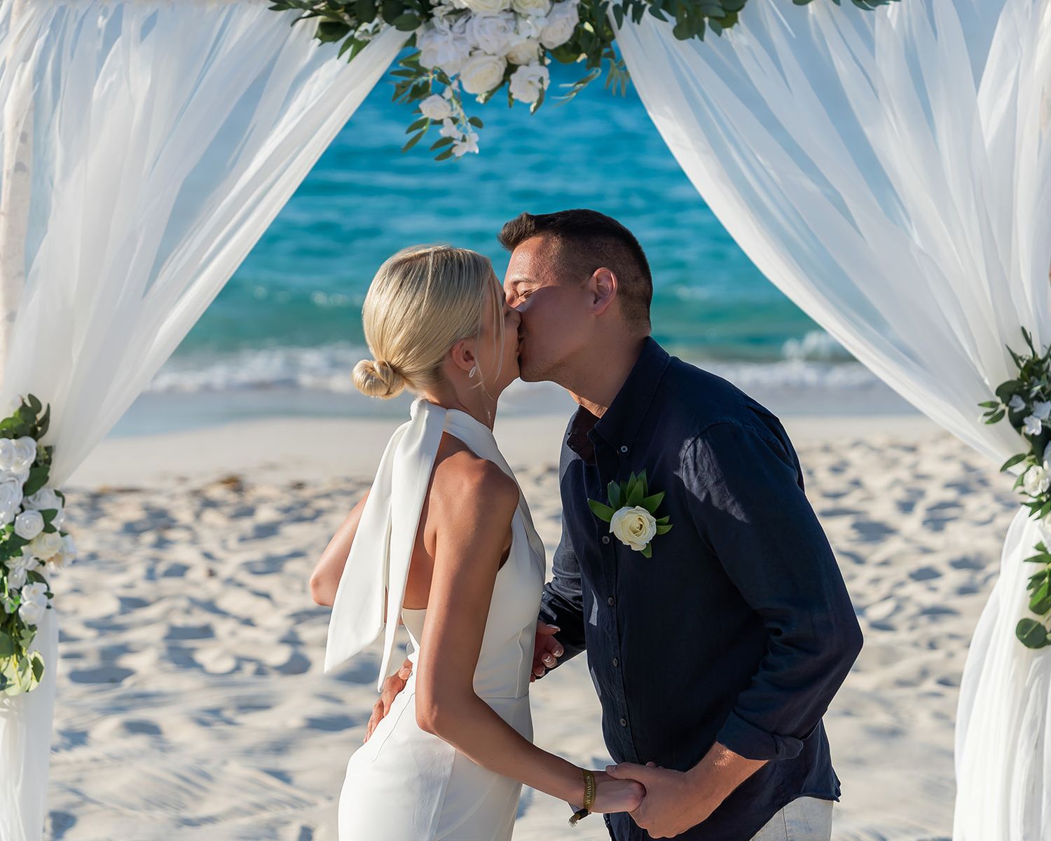 Couple sharing first kiss at beach wedding ceremony under white floral arch with turquoise ocean backdrop.
