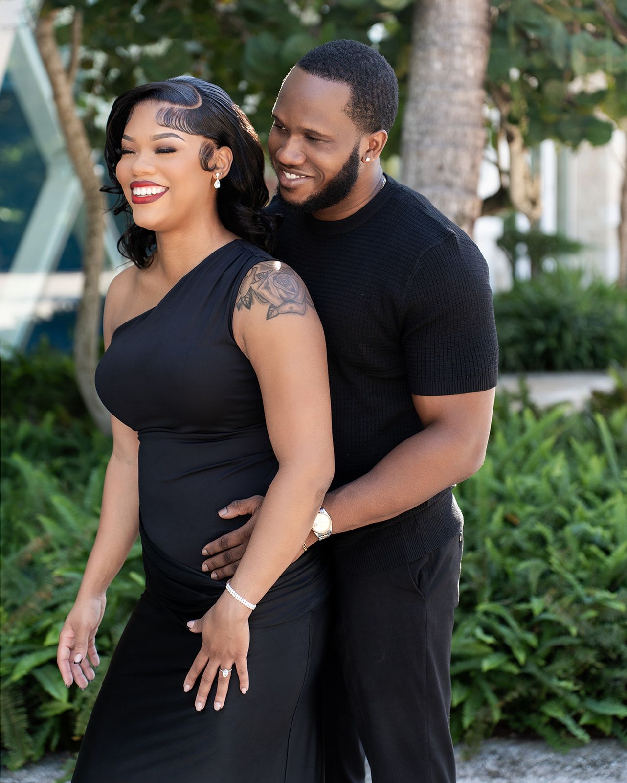 Couple in matching black outfits posing outdoors, man embracing pregnant woman from behind near lush greenery.