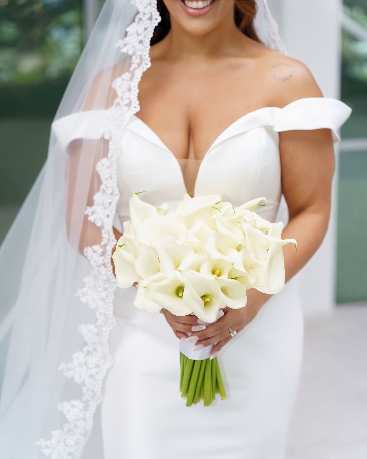 Smiling bride in off-shoulder white gown and lace veil holding elegant white calla lily bouquet on wedding day.