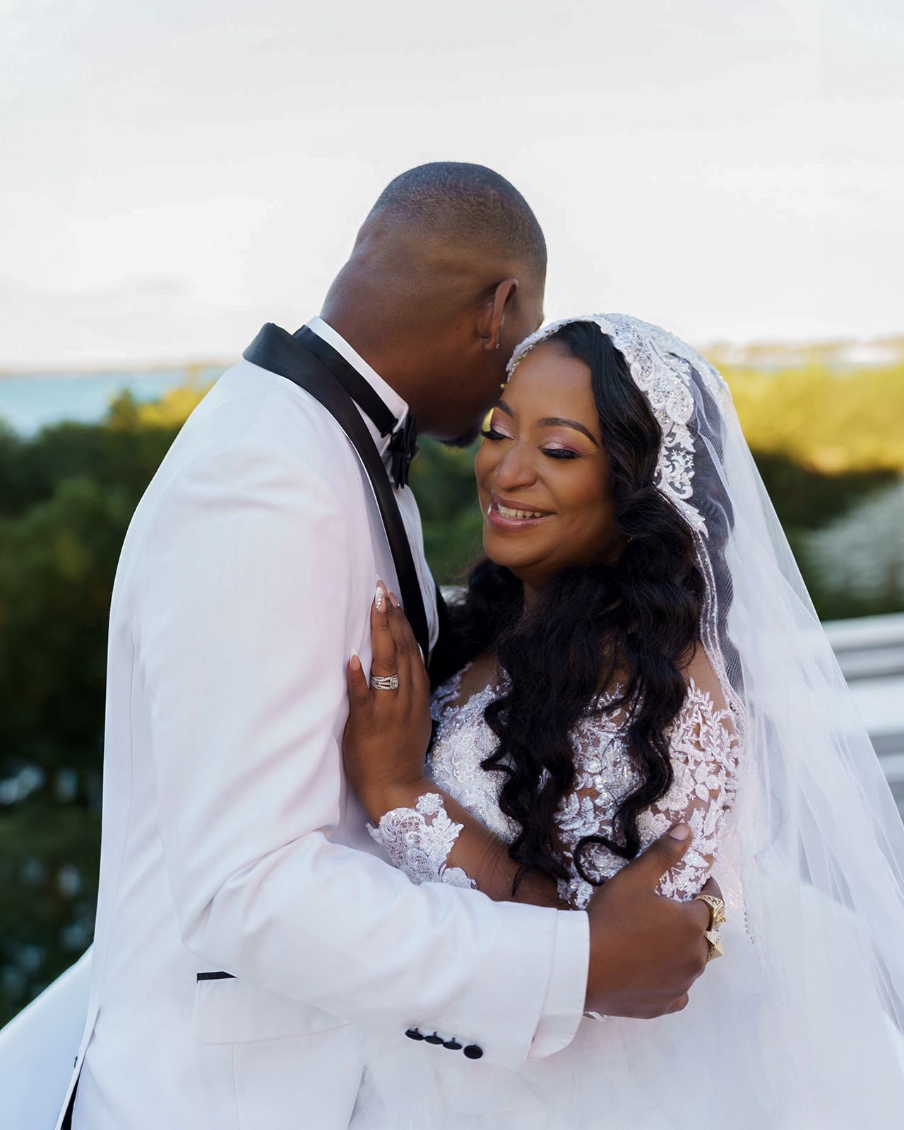 A bride and groom share a romantic embrace outdoors, with the bride in a lace gown and veil, and groom in a white tuxedo.
