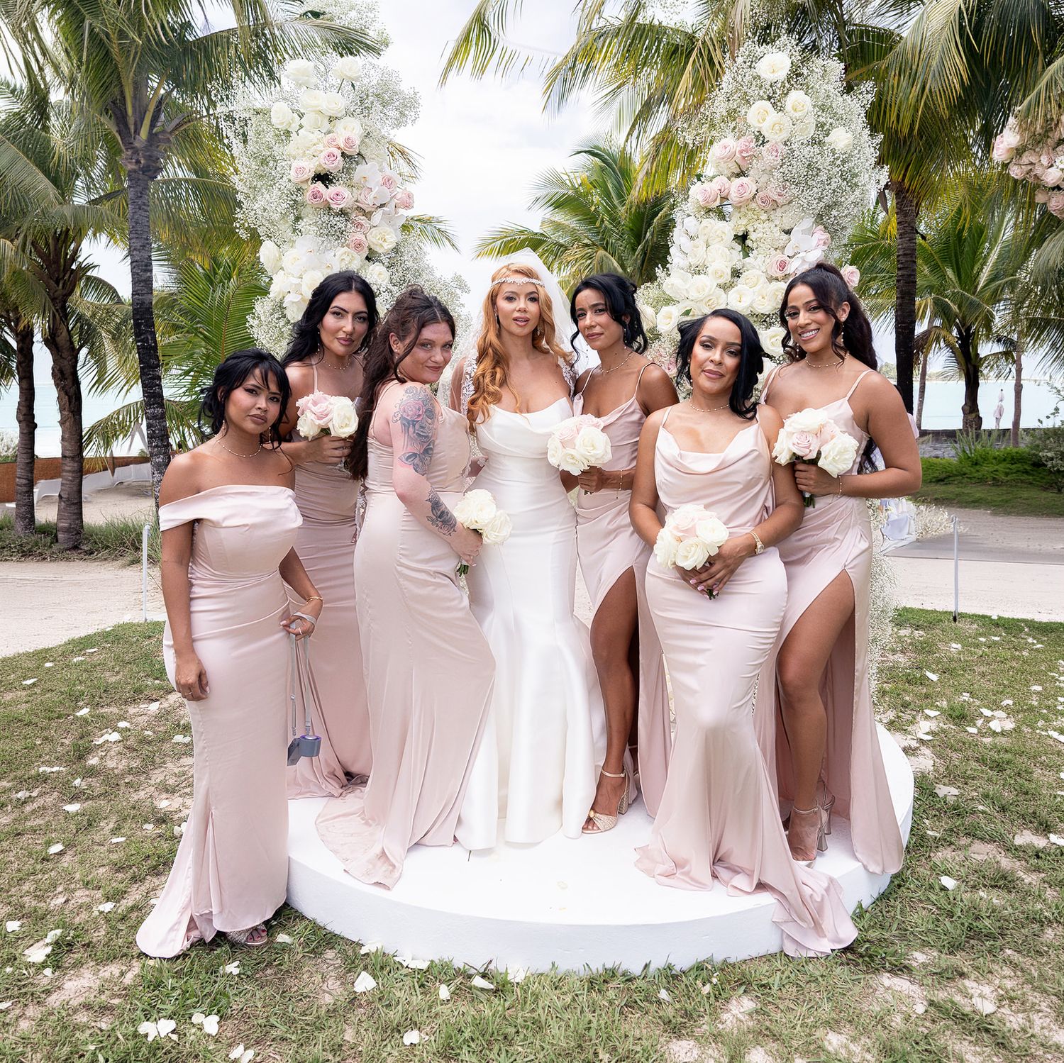 Bride in white gown poses with six bridesmaids in blush pink dresses outdoors near palm trees and floral arch.