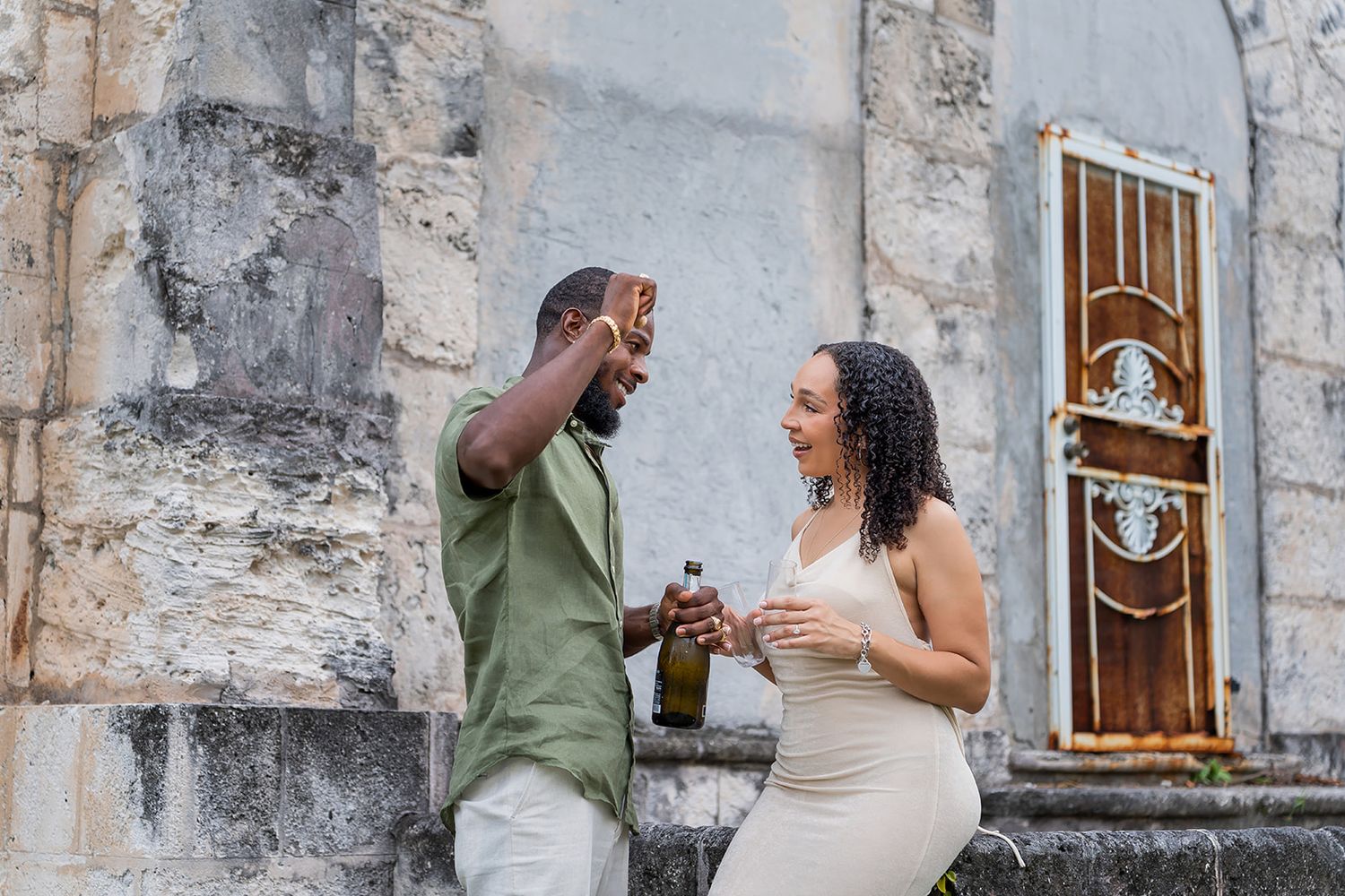 Couple celebrating outdoors near stone building, clinking beer bottles and smiling joyfully.
