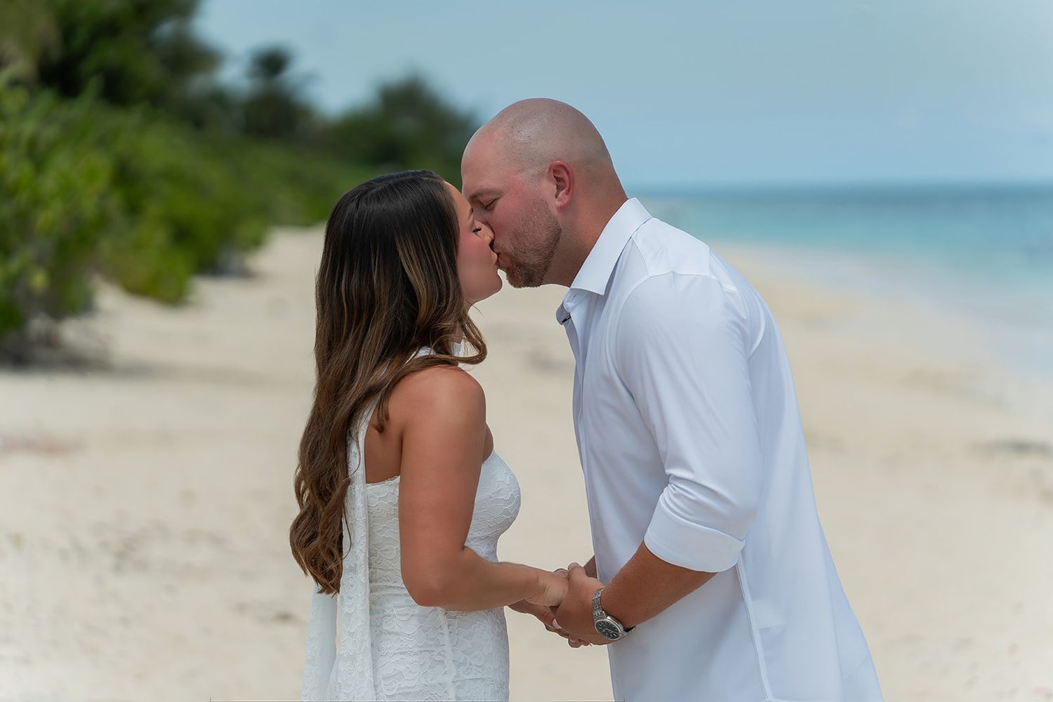 Couple sharing a romantic kiss on a tropical white sand beach, dressed in white outfits with turquoise ocean in background.