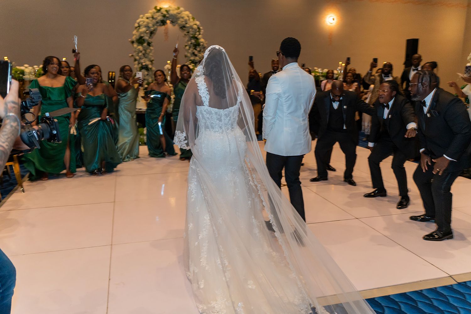 Bride in white gown and veil walks with groom past cheering guests at elegant wedding reception.