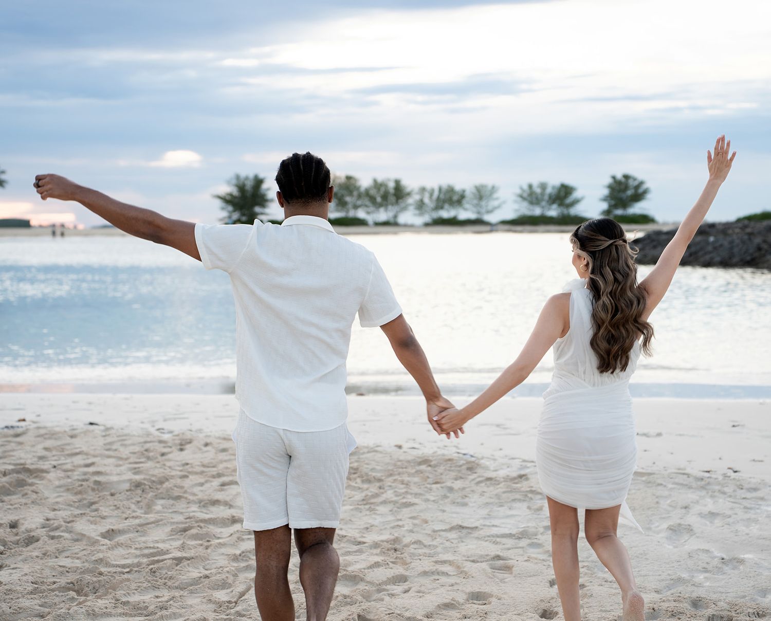 Couple in white outfits holding hands on a sandy beach, arms raised joyfully with ocean and trees in background.