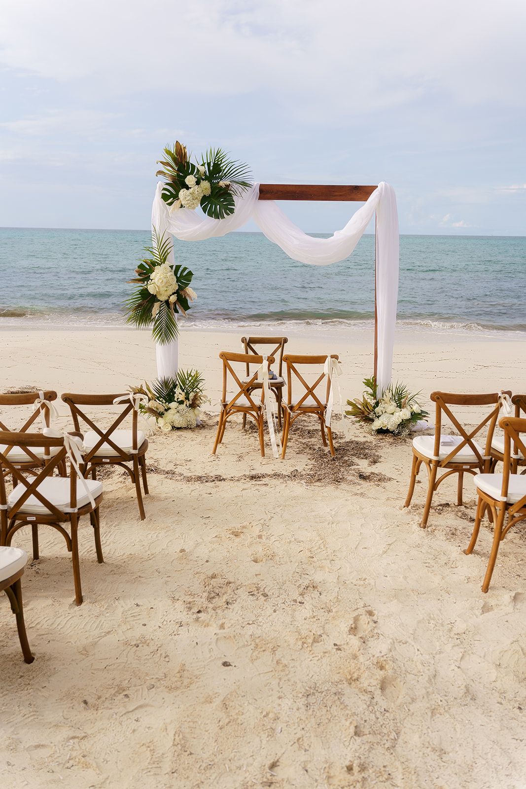 Tropical beach wedding ceremony setup with wooden arch, white draping, floral arrangements, and cross-back chairs on sandy shore.