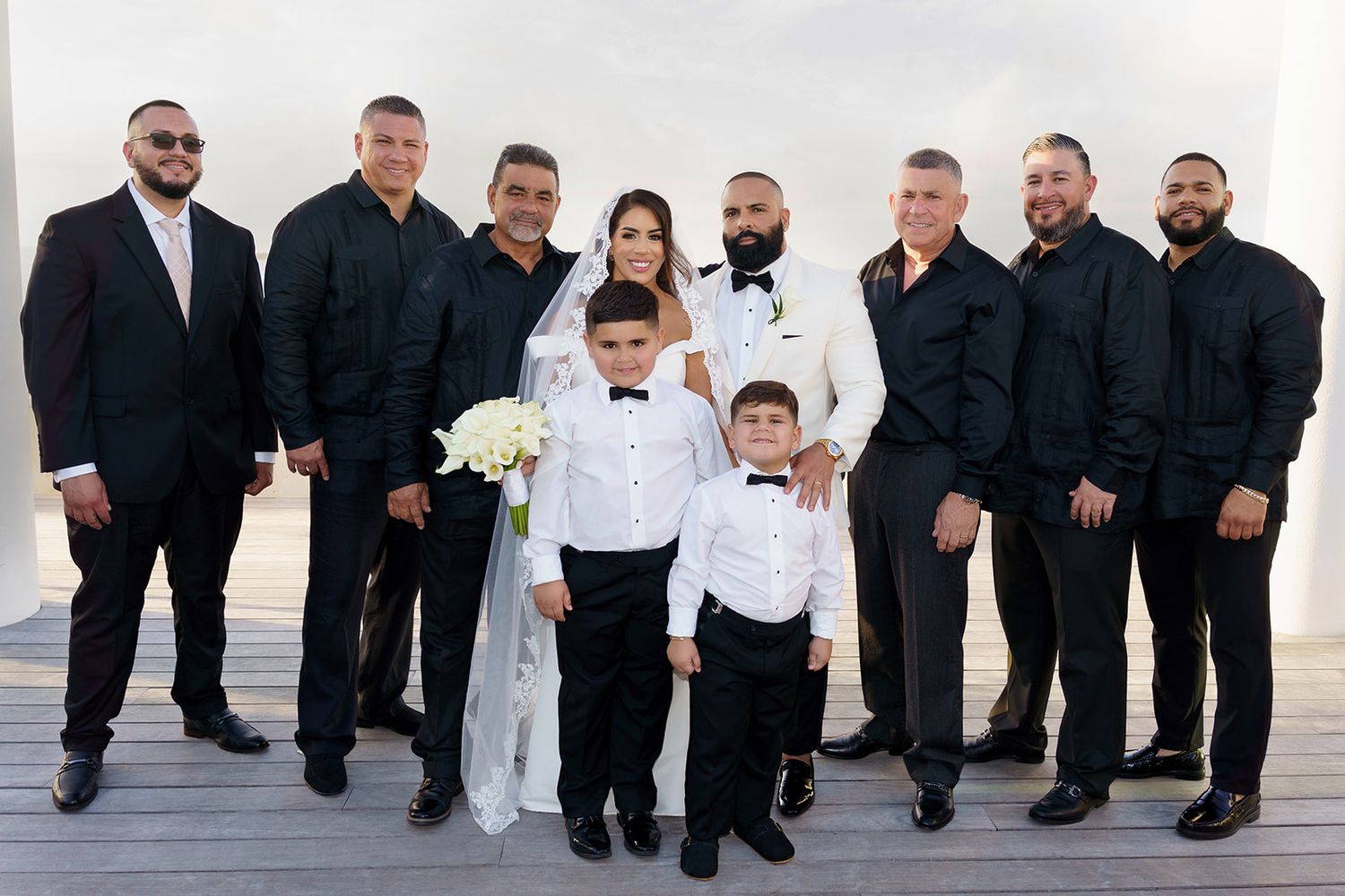 A bride and groom pose with groomsmen in black suits and a young ring bearer at an outdoor wedding venue.