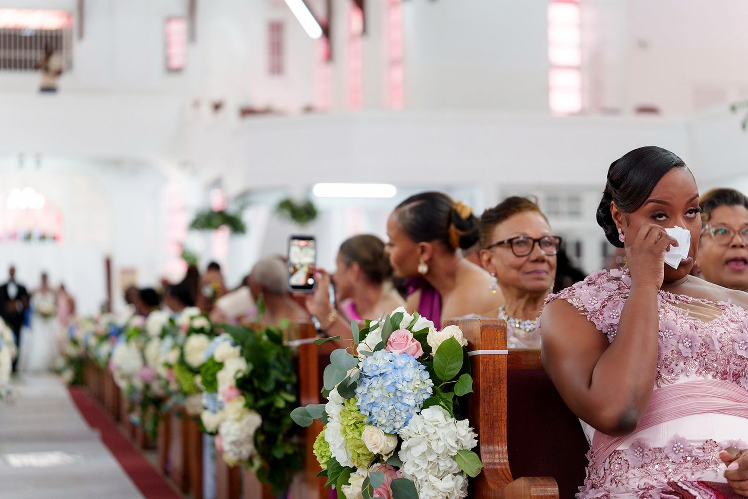 Emotional wedding guests seated in church pews adorned with white and blue floral arrangements during a ceremony.