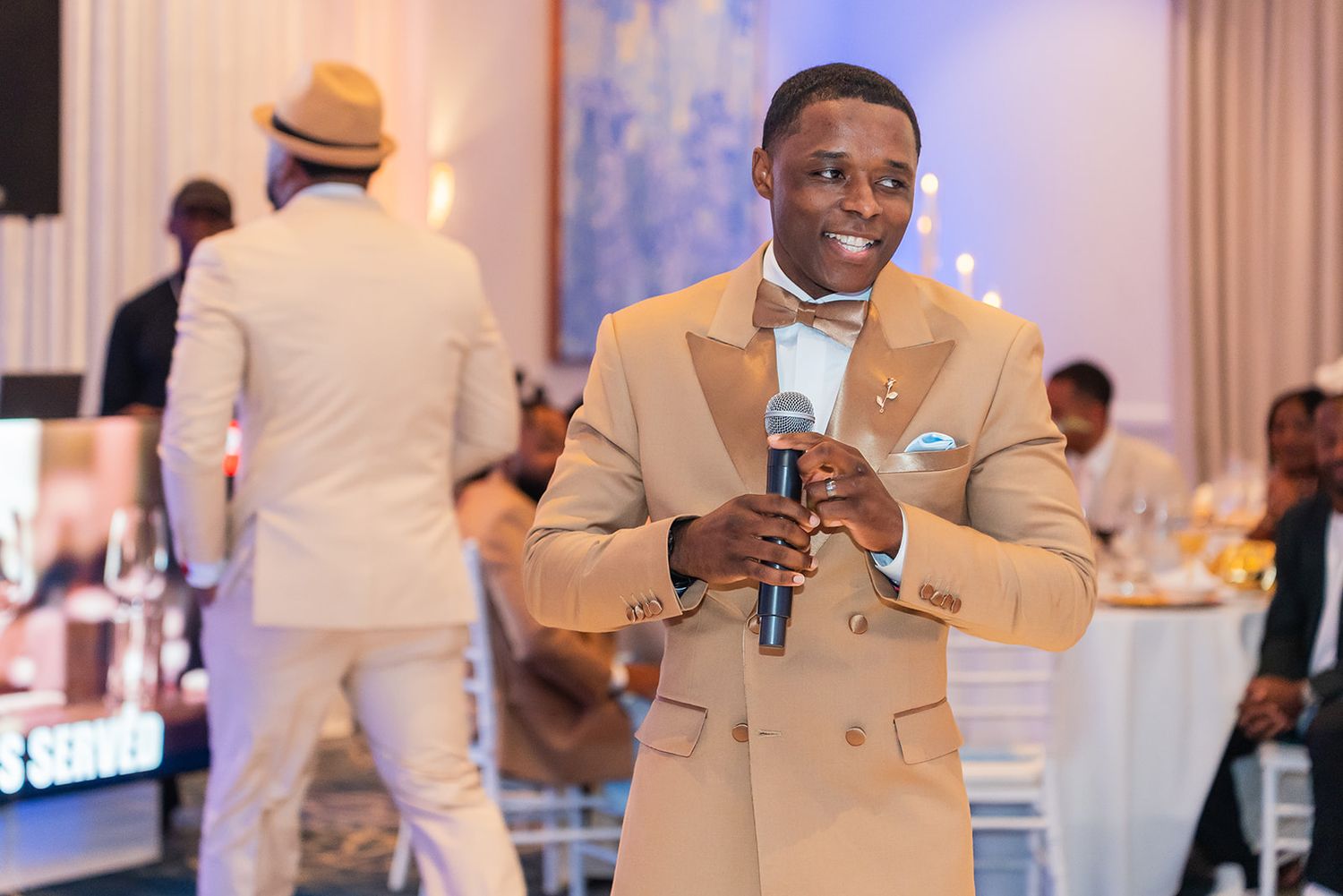 Elegant man in beige suit holding microphone at formal event, smiling confidently at indoor reception venue.
