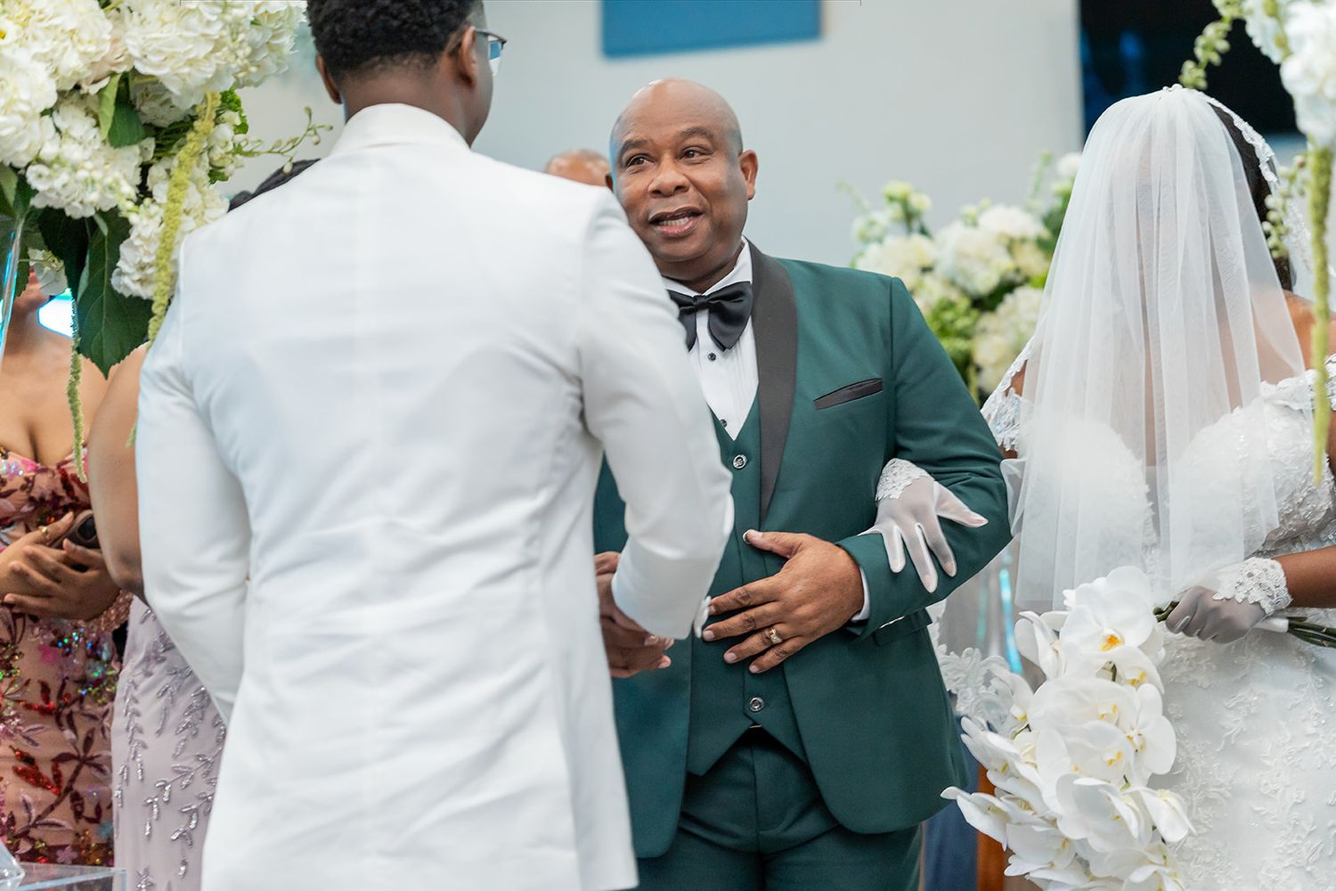 A groom in a teal suit greets a guest at a wedding ceremony with white floral arrangements in the background.
