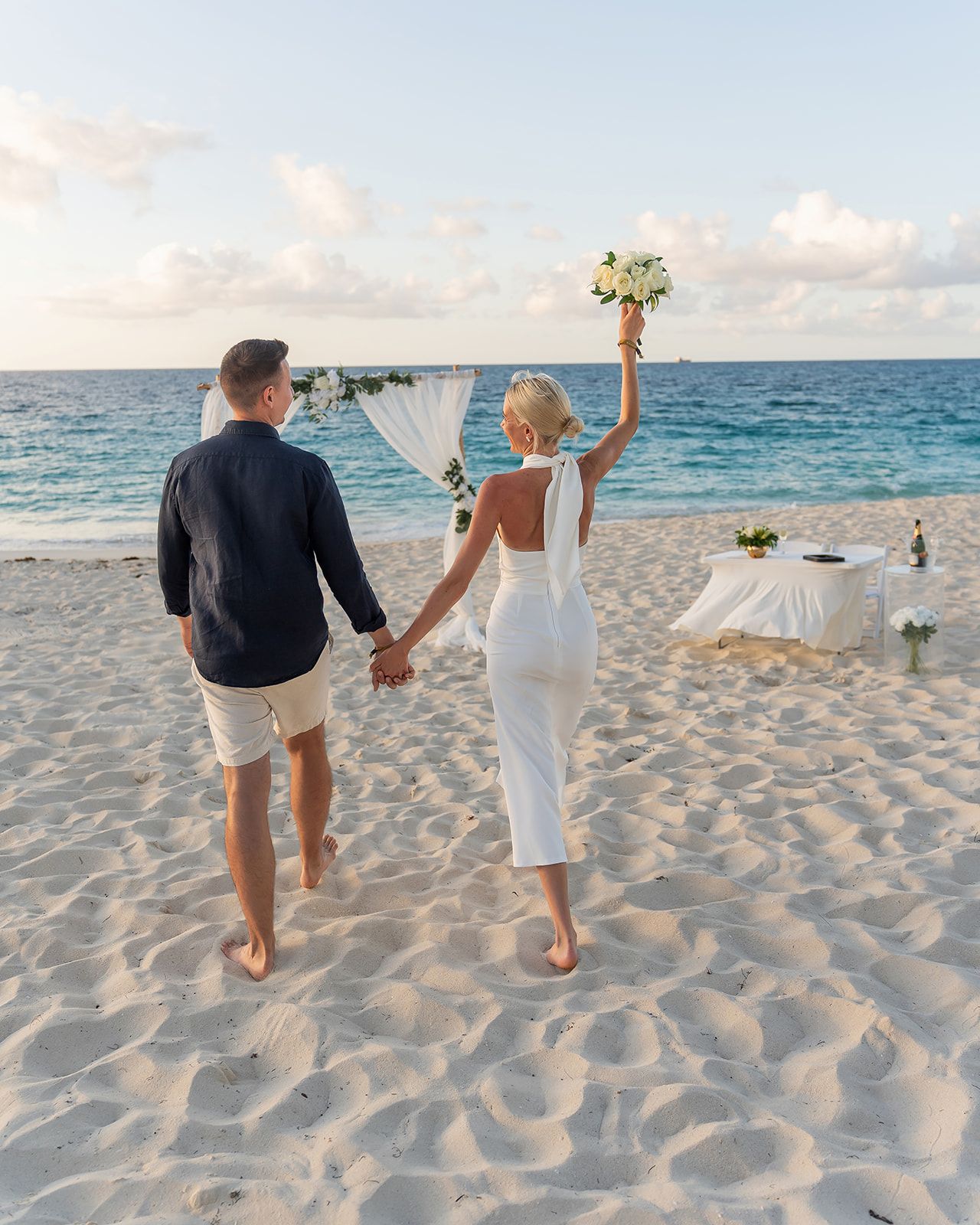 Couple walking hand-in-hand on a sandy beach, bride in white dress raising bouquet, ocean and wedding arch in background.