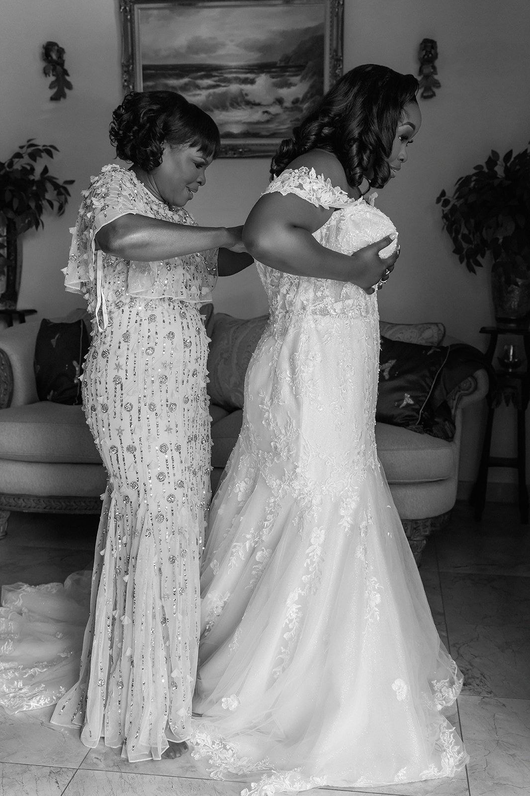 Black and white photo of a bride in lace gown being helped by a woman in beaded dress during wedding preparations.