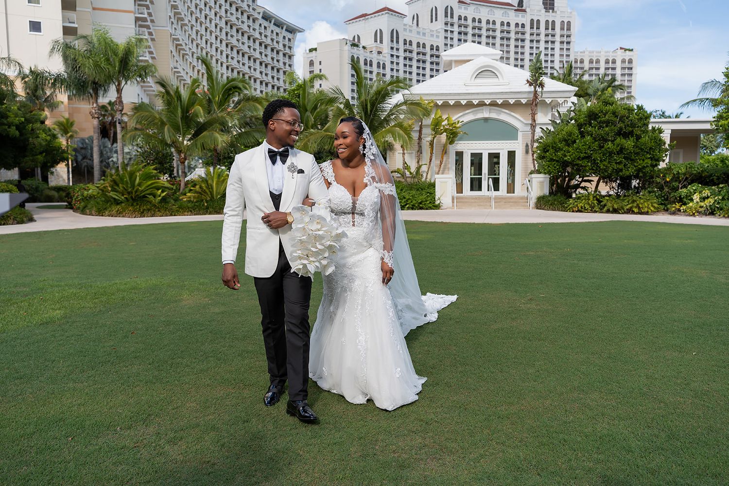 Bride and groom posing on lush green lawn at elegant resort wedding venue with palm trees and white chapel.