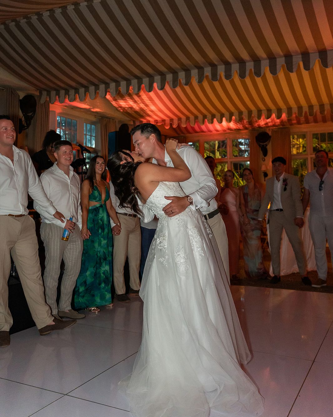 Bride and groom share first dance kiss at elegant wedding reception with guests watching under striped tent canopy.
