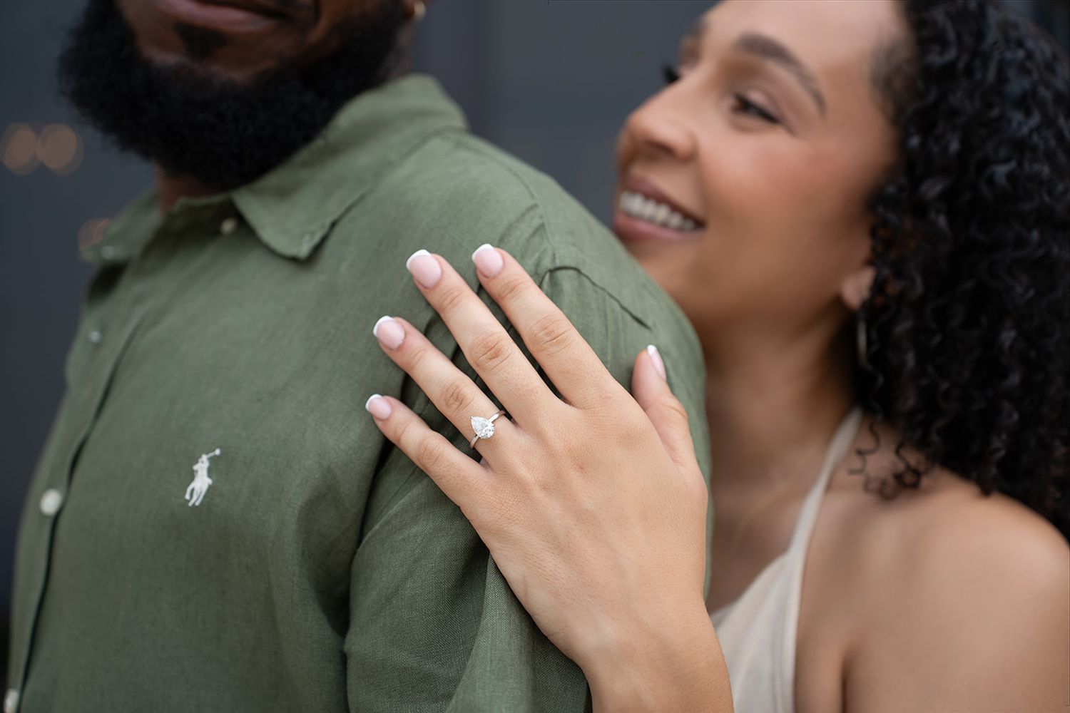 Close-up of woman showing off diamond engagement ring while smiling and resting hand on partner's green shirt.