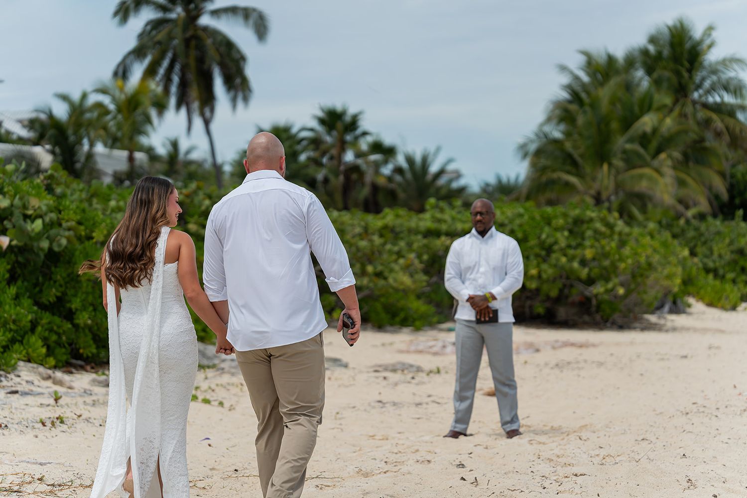 Couple in white attire walks hand-in-hand on a tropical beach toward an officiant during an outdoor wedding ceremony.