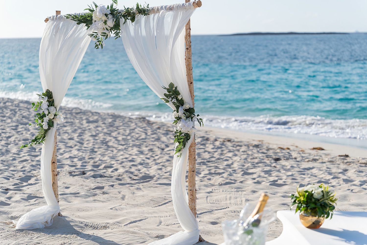White draped wedding arch decorated with greenery on a sandy beach with turquoise ocean backdrop.