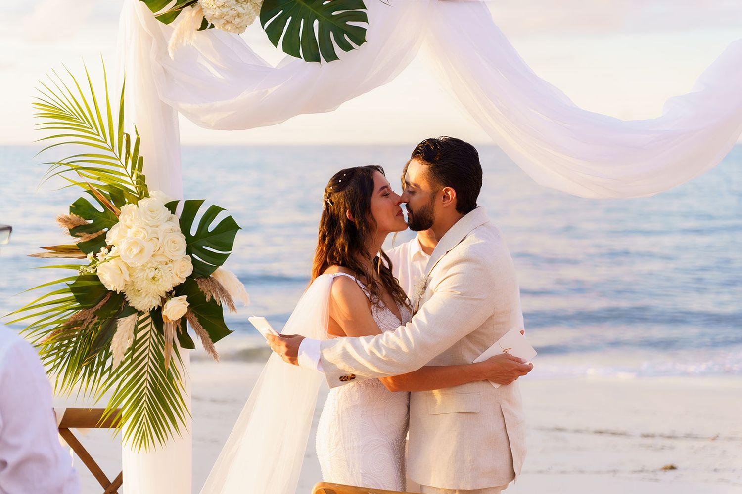 Couple sharing first kiss at tropical beach wedding ceremony with white draping and palm leaf floral arch by the ocean.