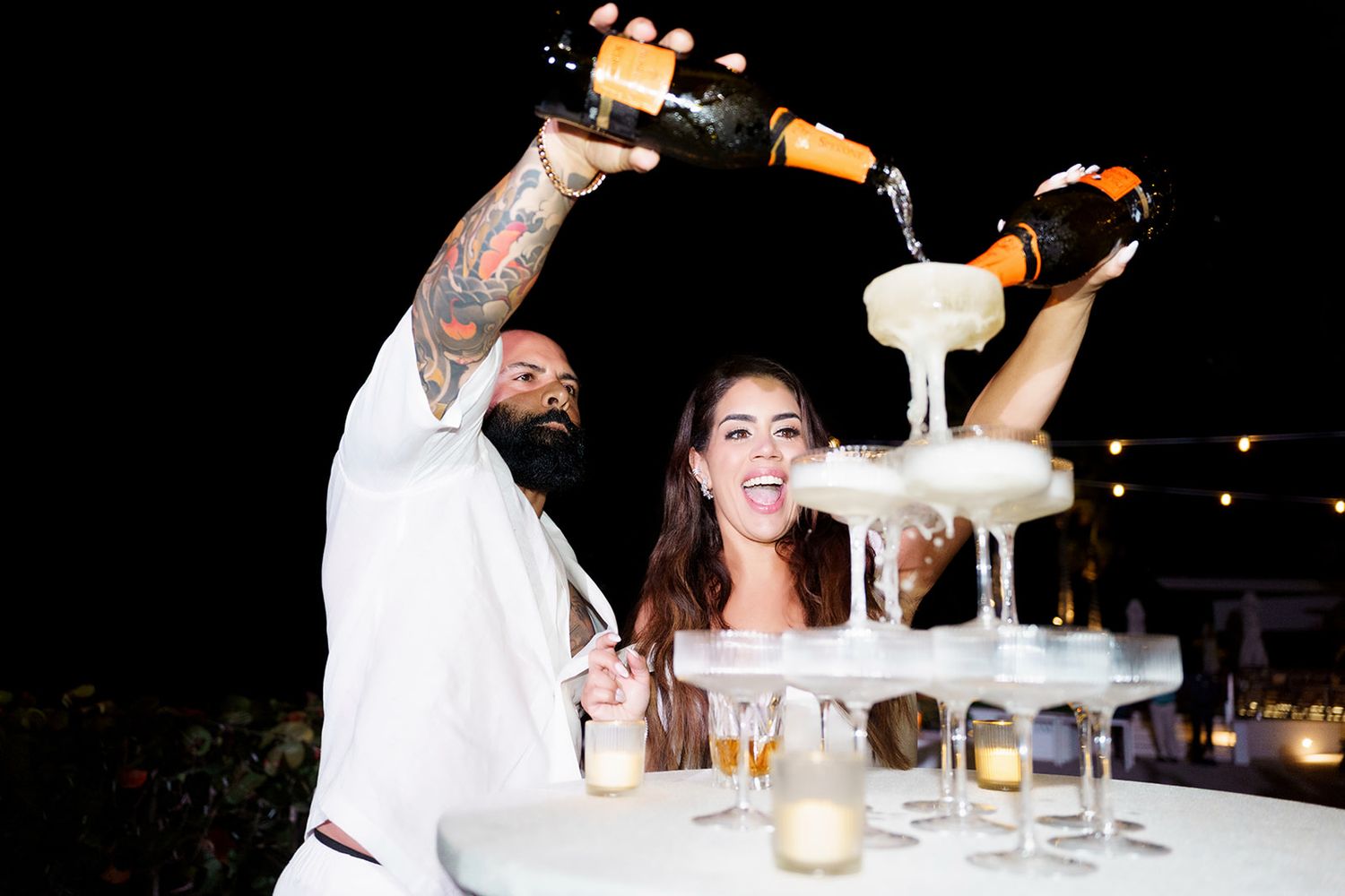 Couple pouring champagne into a tower of glasses at a wedding reception at night.