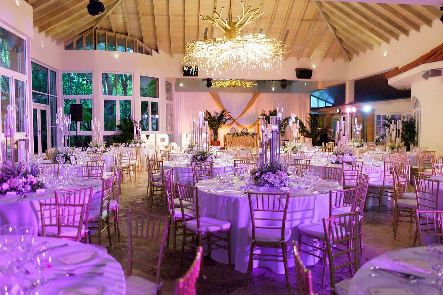 Elegant wedding reception hall with gold Chiavari chairs, white round tables, floral centerpieces, and purple ambient lighting.