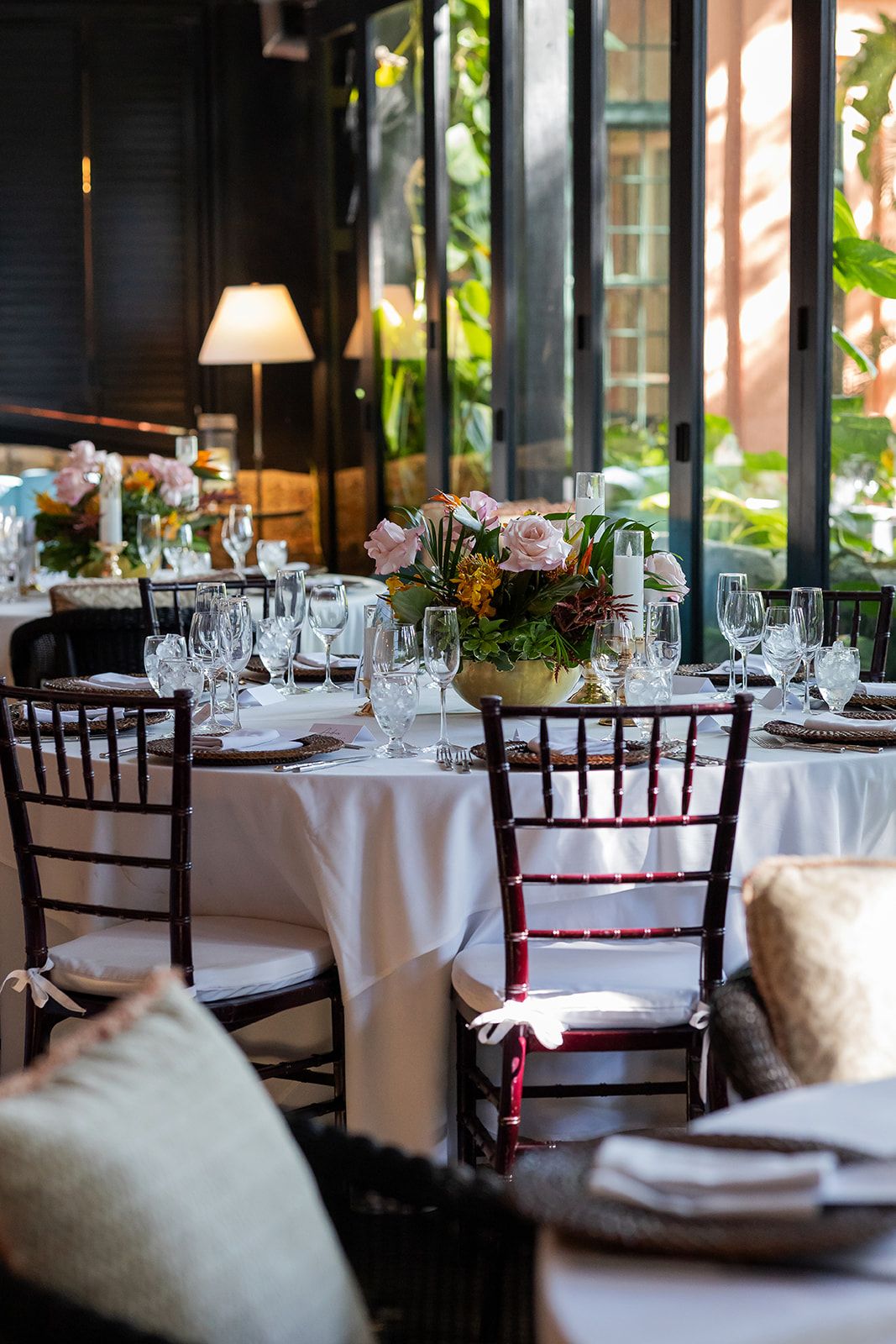 Elegant restaurant dining room with white tablecloths, dark Chiavari chairs, crystal glassware, and floral centerpieces near large windows.