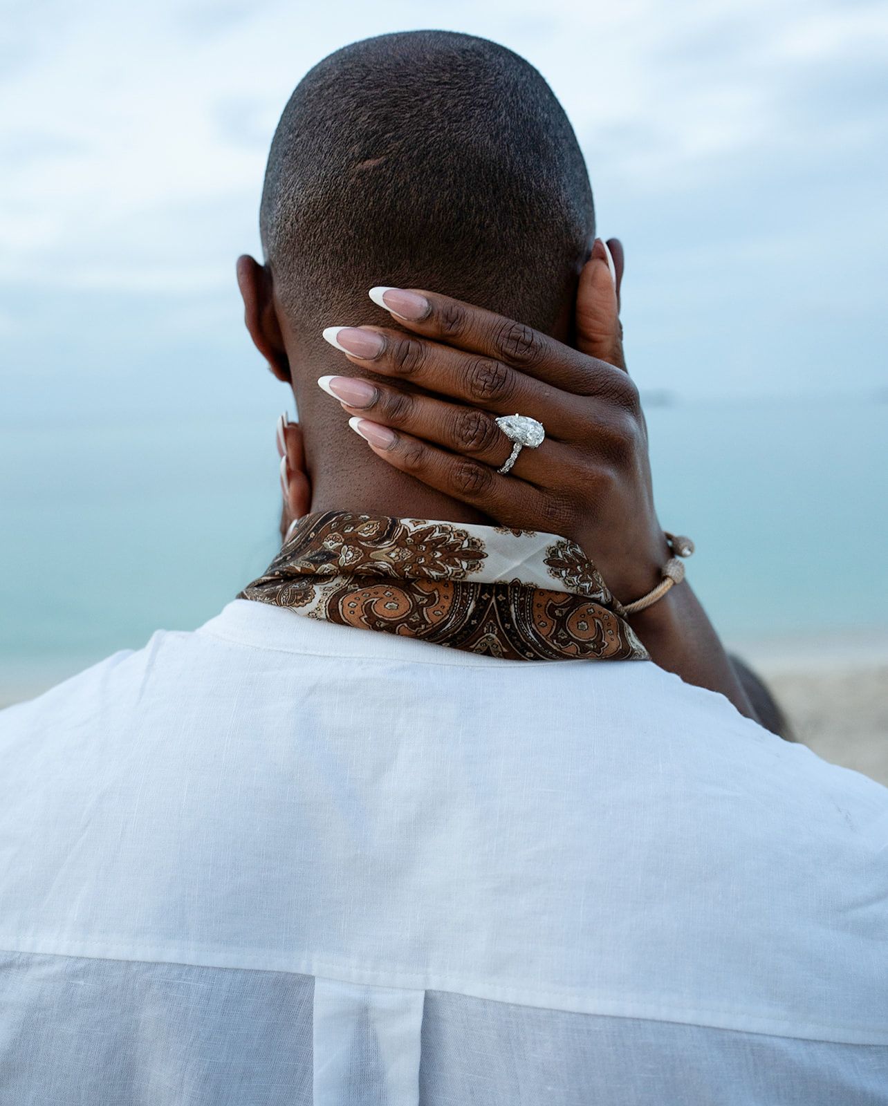 A woman with henna tattoos and a diamond ring places her hand on a man's neck near the ocean.