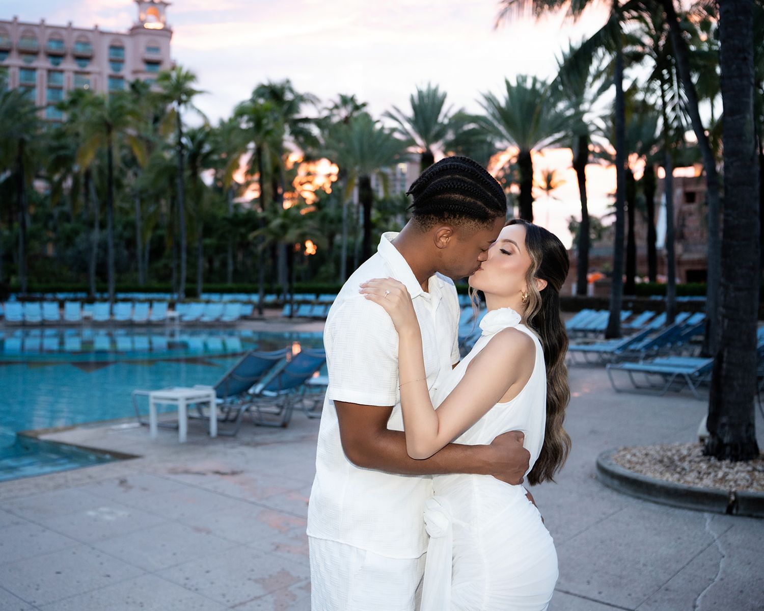 Couple in white outfits sharing a romantic kiss by a resort pool surrounded by palm trees at sunset.