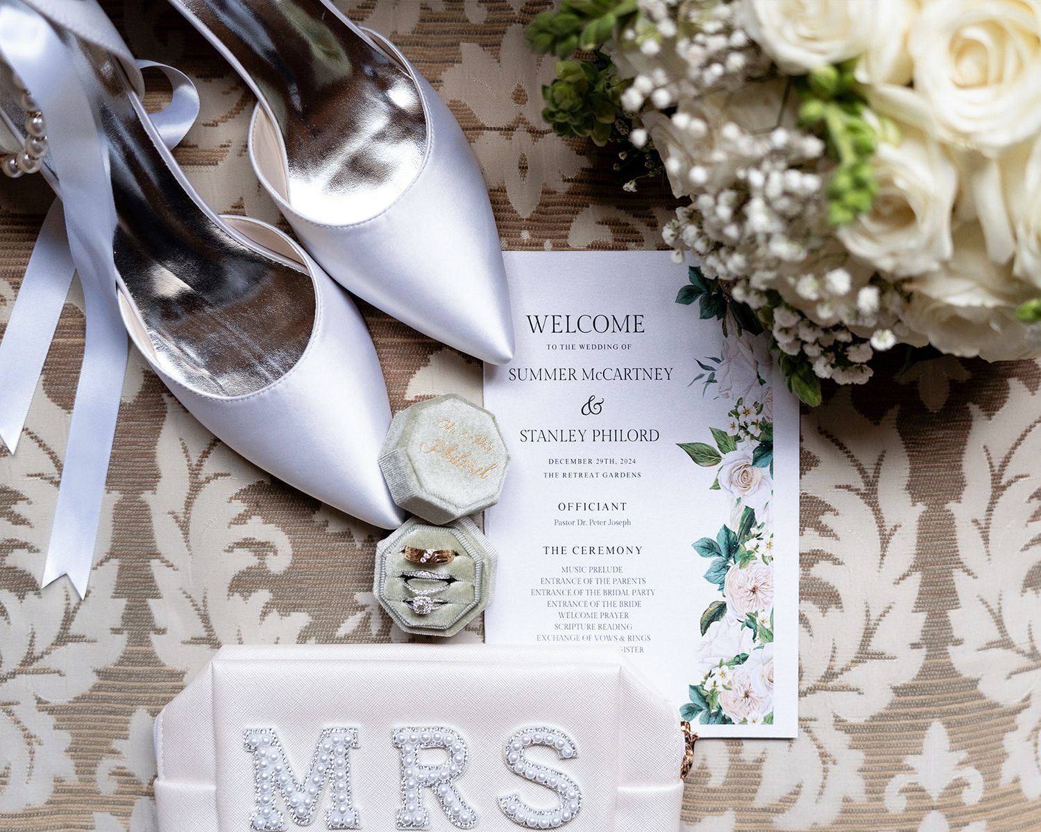 Wedding flat lay with white heels, floral bouquet, program, ring box, and MRS sign on elegant fabric.