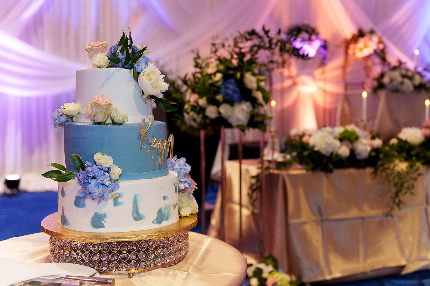 Elegant three-tier blue and white wedding cake adorned with flowers and gold details at a reception venue.