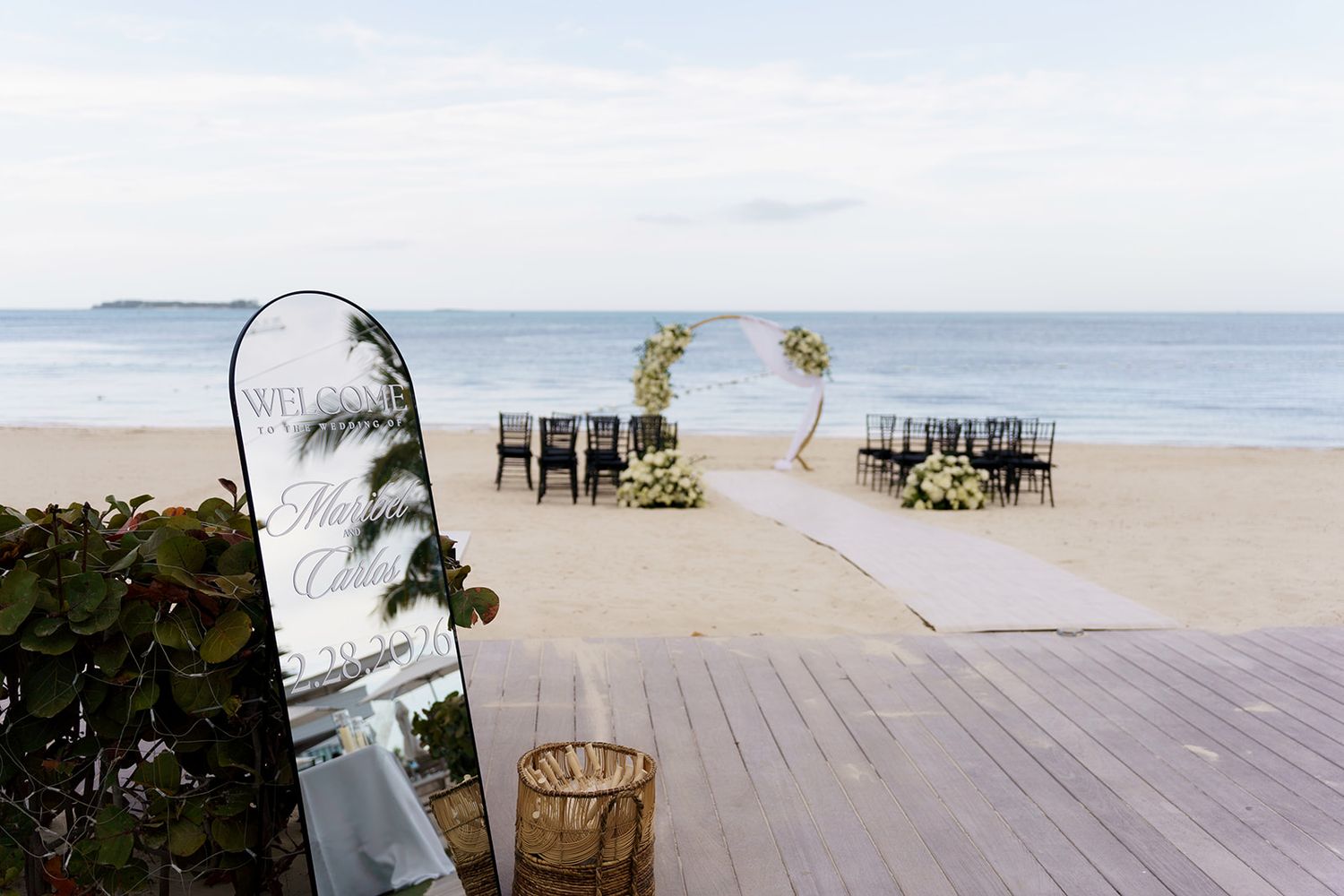 Elegant beach wedding setup with black chairs, floral arch, and aisle runner on sandy shore with ocean backdrop.