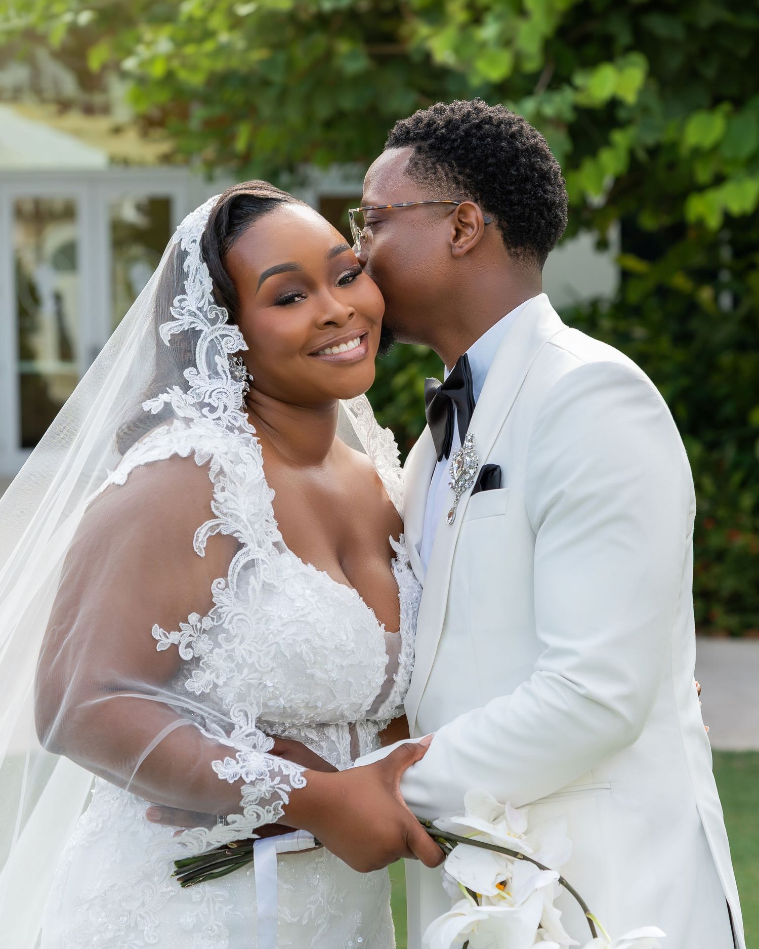 A joyful bride in lace wedding gown and veil holds flowers as groom in white suit kisses her cheek outdoors.