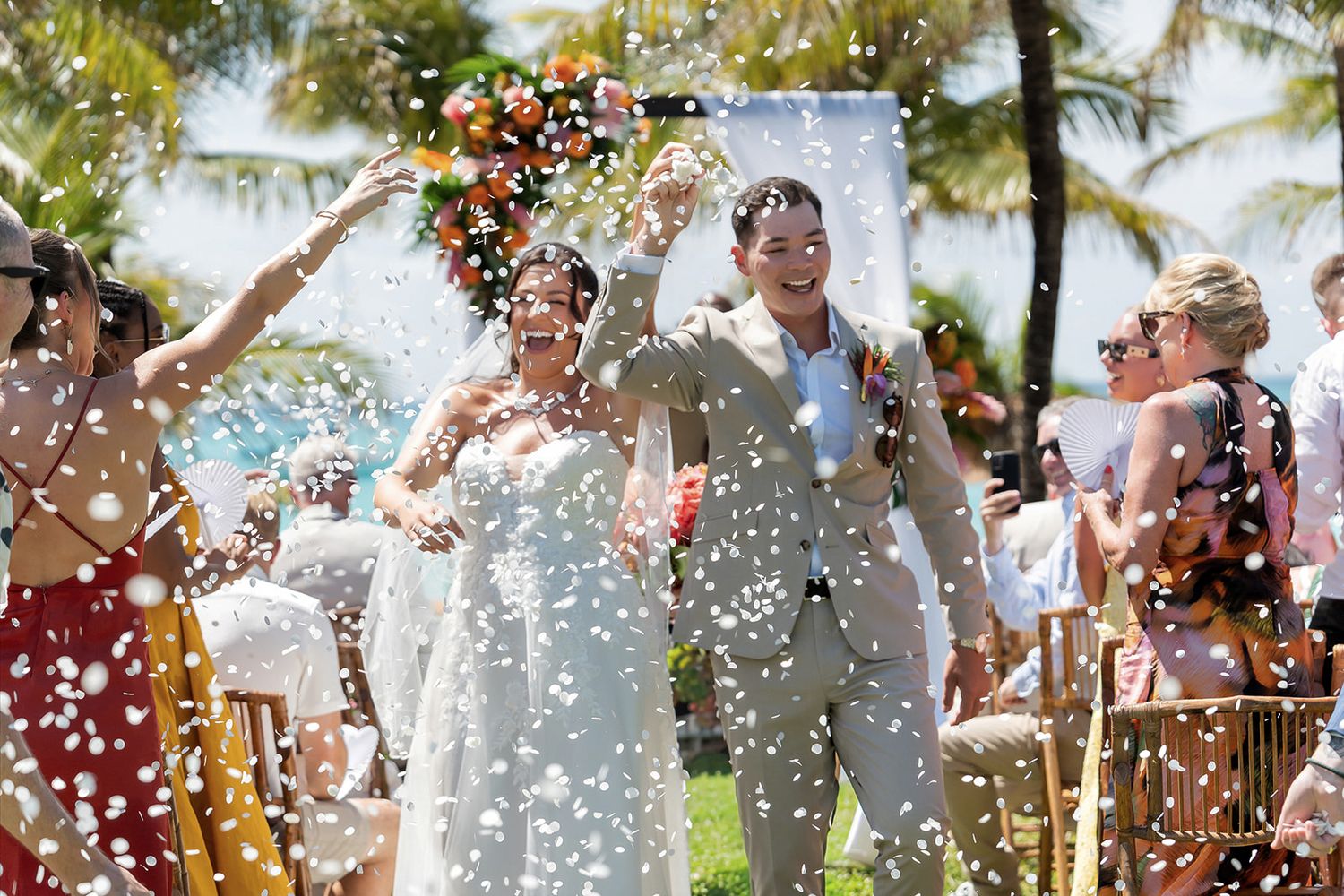 Bride and groom walk through confetti shower at tropical outdoor wedding ceremony with palm trees.