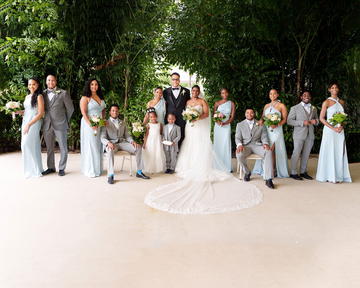 Wedding party poses outdoors in lush garden setting, bridesmaids in blue gowns and groomsmen in gray suits flanking bride and groom.