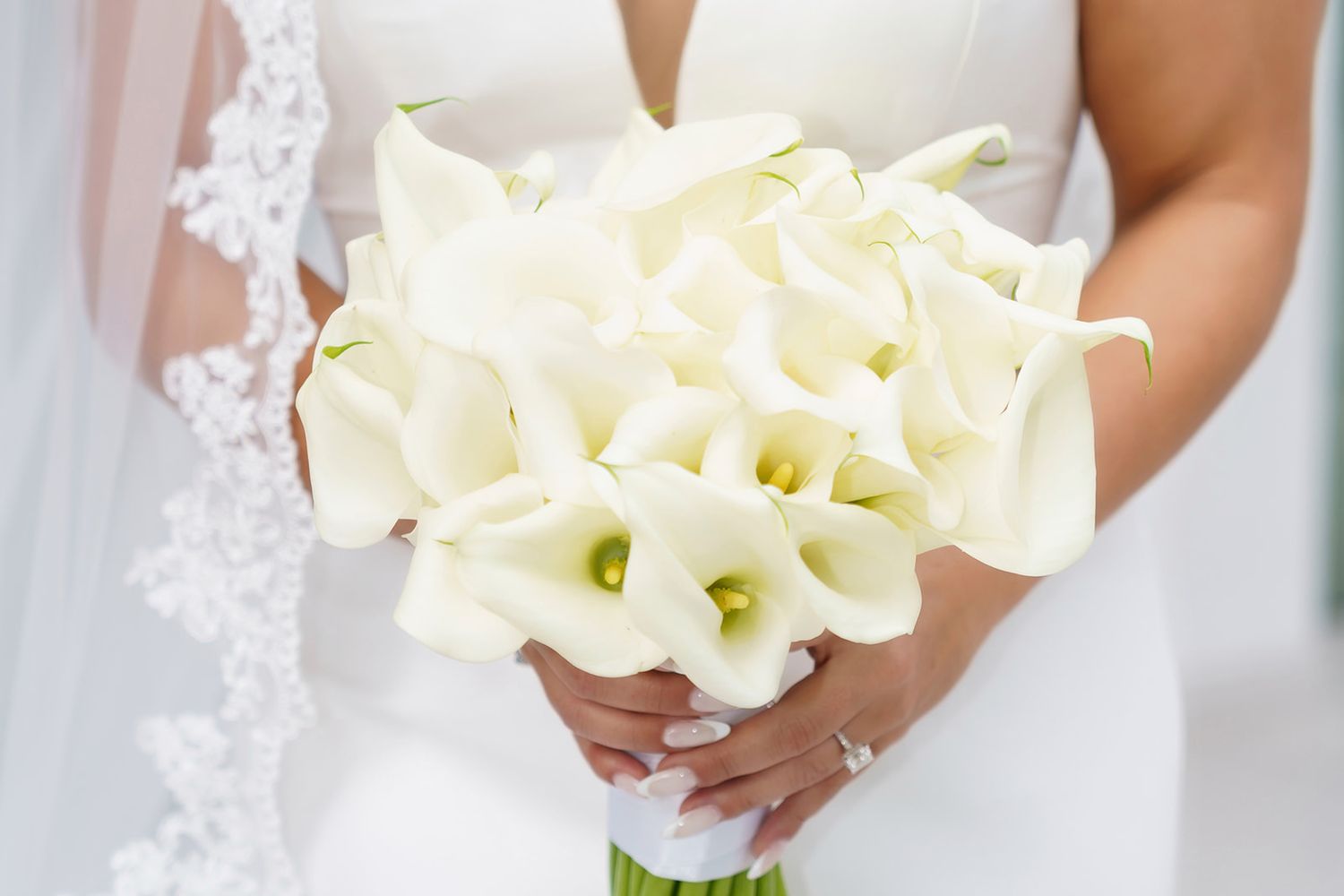 Bride in white wedding dress holding elegant bouquet of white calla lilies, showcasing diamond ring on manicured hands.