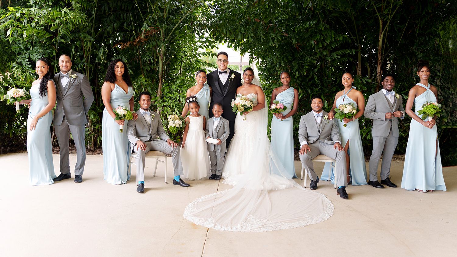 Wedding party poses together outdoors, bridesmaids in light blue gowns and groomsmen in gray suits.