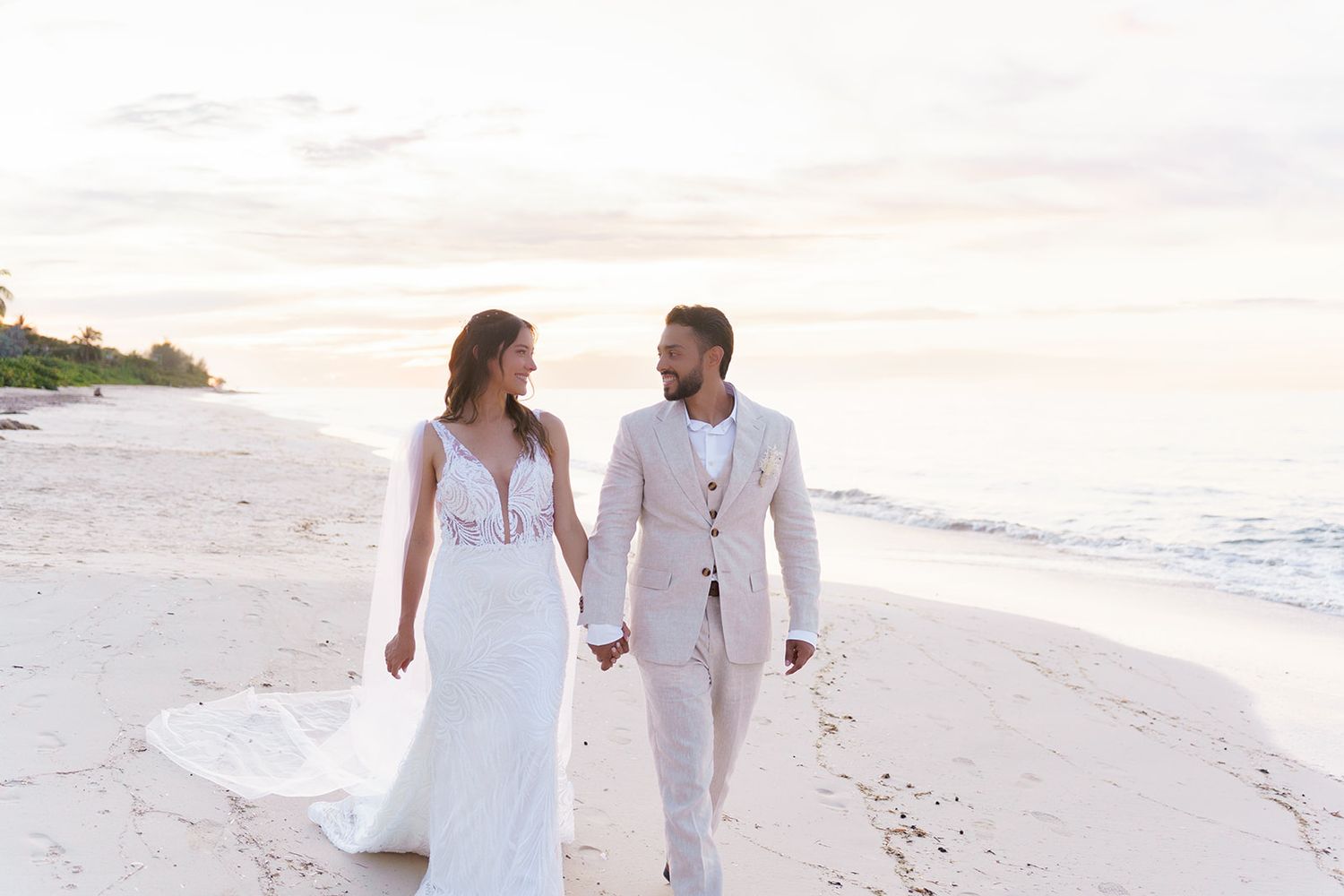 Bride and groom walk hand-in-hand on a sandy beach at sunset, wearing elegant white wedding attire.