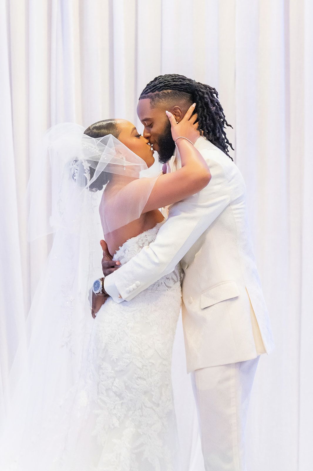Bride and groom sharing first kiss at wedding ceremony, both dressed in white, against draped white backdrop.