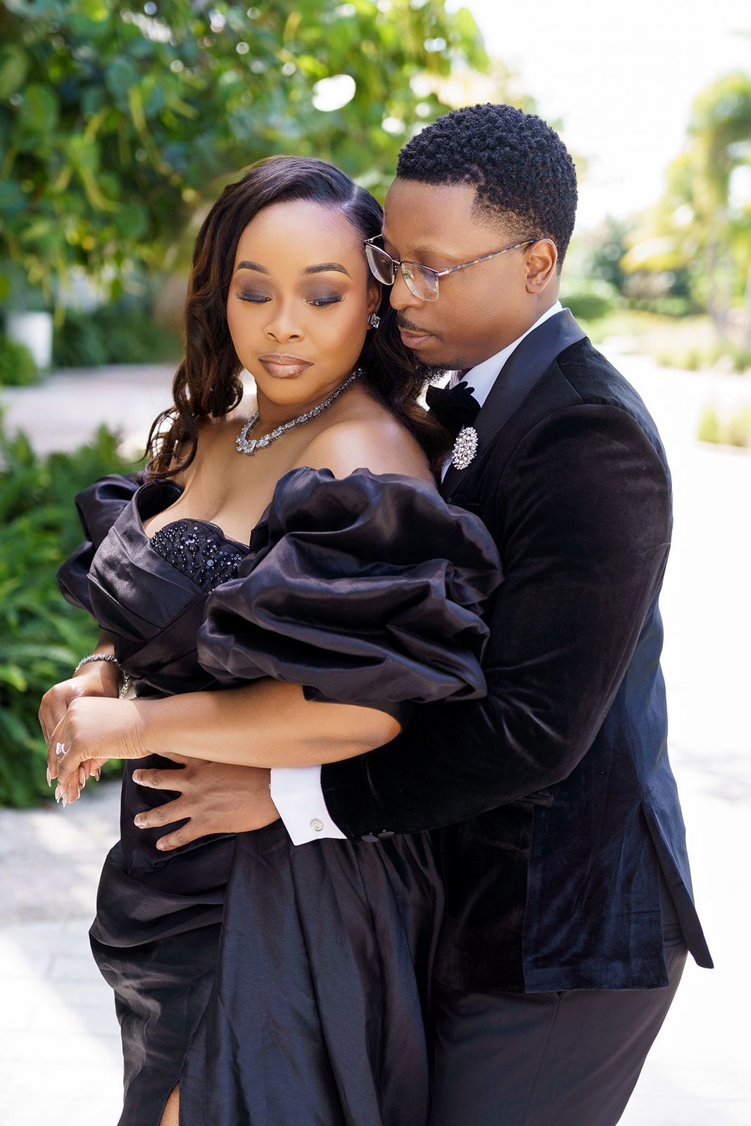 Elegant Black couple embracing outdoors, woman in off-shoulder black gown, man in velvet suit with glasses.