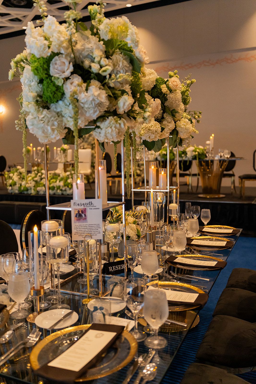Elegant wedding reception table with tall white floral centerpieces, gold accents, and candles.