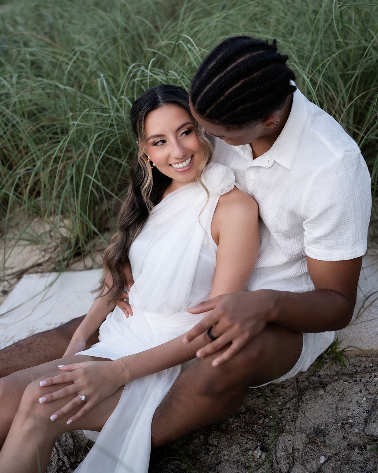 Couple in white outfits sitting on sand dunes, woman smiling with engagement ring, man kissing her cheek.