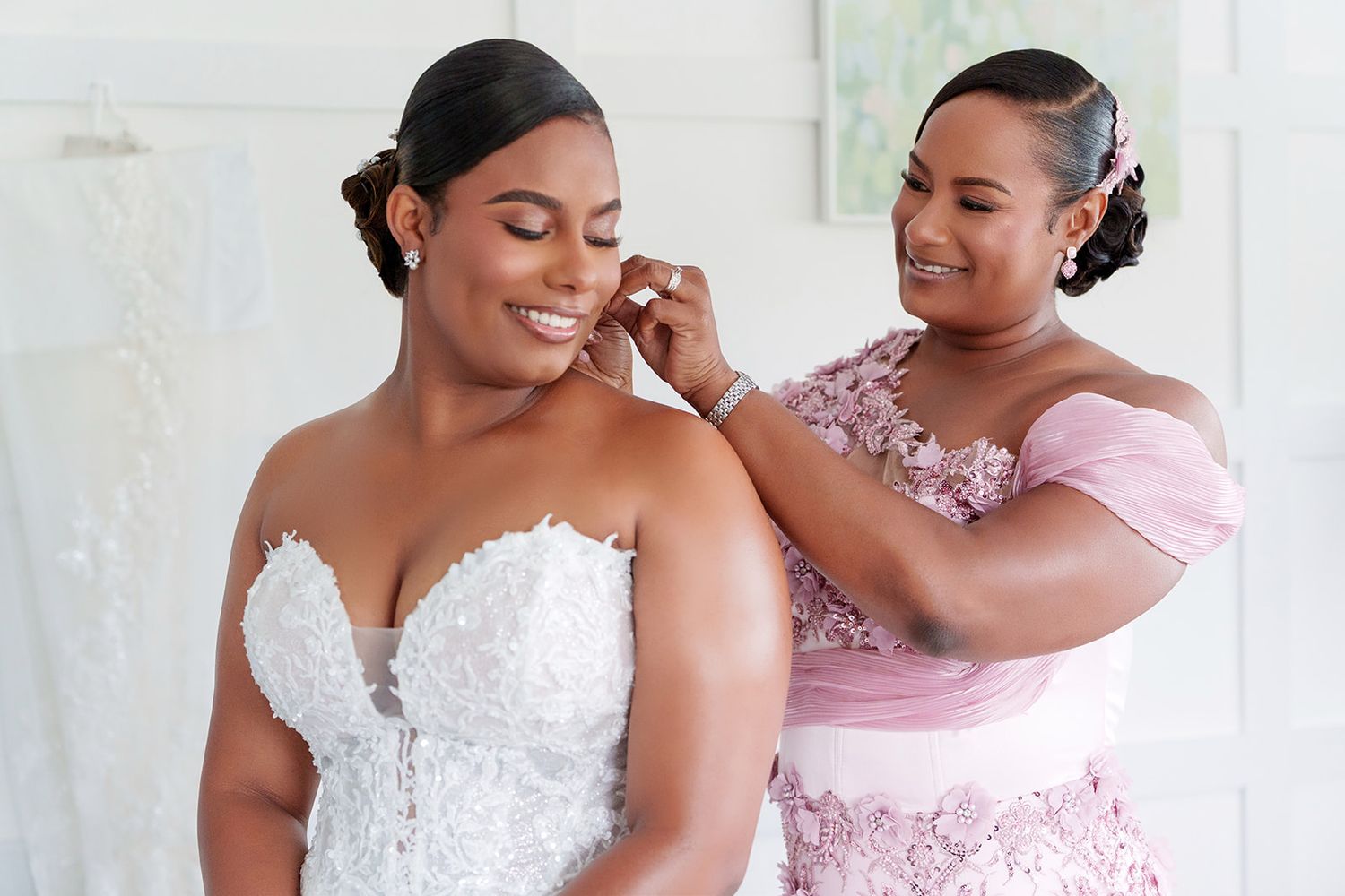 Bridesmaid helping bride put on earrings while smiling in a bright wedding preparation room.